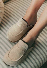 Beige Filzhausschuhe mit Lederakzenten, runder Zehenform und schwarzem Innenfutter. An den Füßen getragen, liegen auf einer gestreiften Oberfläche.