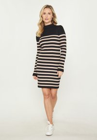 Gestreiftes Strickkleid in Schwarz und Beige mit Stehkragen und langen Ärmeln, kombiniert mit weißen Sneakers. Figurbetonter Schnitt.