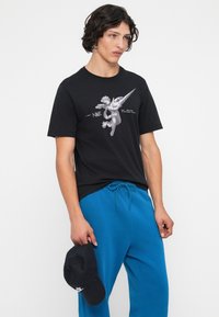 Mlad moški nosi črno majico s kratkimi rokavi z grafiko cheruba in modre hlače, v desni roki drži črno kapo Nike.