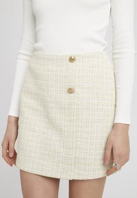 Mini-jupe en tweed blanc et jaune avec un motif texturé, ornée de deux boutons dorés sur le devant. Conception droite avec une finition lisse.