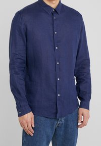 Uomo che indossa una camicia a maniche lunghe blu navy con bottoni abbinata a jeans blu su uno sfondo grigio uniforme.