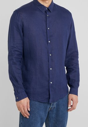 Uomo che indossa una camicia a maniche lunghe blu navy con bottoni abbinata a jeans blu su uno sfondo grigio uniforme.