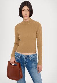 Un haut en tricot côtelé beige avec un col montant et des manches longues, associé à un jean en denim bleu. Tenant un sac à main en cuir marron de forme incurvée.