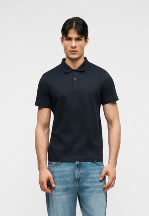 Jonge man met donker haar, gekleed in een zwart poloshirt met korte mouwen en lichtblauwe spijkerbroek, staand tegen een effen lichte achtergrond.