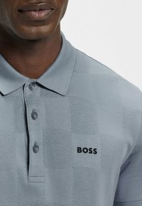 Szary polo z kratkowanym wzorem, z trzema guzikami przy kołnierzyku i czarnym logo "BOSS" na klatce piersiowej. Zbliżenie, bez widoku całej sylwetki.