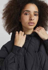 Veste noire matelassée avec un col montant, des boutons de fermeture et des manches bouffantes, mettant en valeur un tissu texturé et un design sans couture.