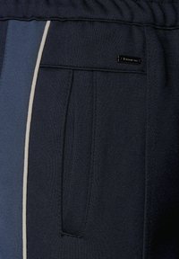 Marineblaue Sporthosen mit horizontalen Farbstreifen in dunklem und hellem Blau, einer Seitentasche und einem kleinen Markenschild.