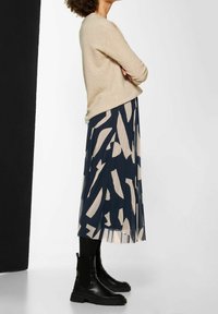 Maglione beige a maglia abbinato a una gonna plissettata blu navy con motivi astratti in beige chiaro, abbinato a stivali neri alla caviglia.