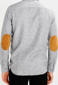 Šedá vlnená košile s tečkovanou texturou, s hnědými semišovými záplaty na loktech a knoflíkovými manžetami. Viditelné švy podél lemu a límce.