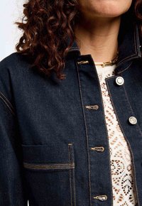 Veste en denim noir avec des coutures contrastées, une poche avant et des boutons en argent ; portée par-dessus un top blanc en dentelle texturée.
