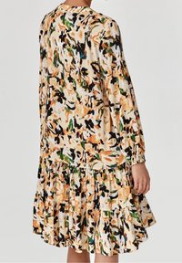 Vestido con estampado floral en crema, que presenta tonos naranjas, negros y verdes. Mangas largas con puños fruncidos, dobladillo con volantes y silueta relajada.