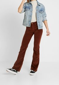 Pantalons en velours côtelé marron évasés, veste en denim et pull léger. Baskets noires avec semelles blanches. Modèle prenant une pose dynamique.