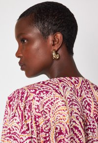 Blusa a fantasia colorata con vortici rosa, arancio e bianco, caratterizzata da un collo arricciato. Orecchino a cerchio dorato che aggiunge dettaglio.