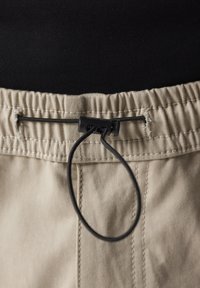 Pantalones beige con cintura elástica y cordón negro. La tela tiene una textura suave y presenta costuras a lo largo de la cintura.
