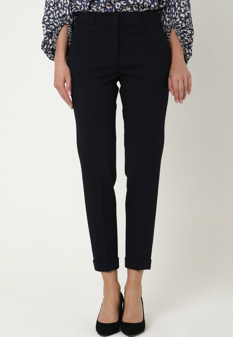 Pantalon noir sur mesure longueur cheville avec ourlets retroussés porté avec des talons noirs et un chemisier bleu à motifs floraux.