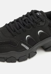 Chaussure de sport noire avec une tige en mesh, semelle en caoutchouc épaisse et accents réfléchissants. Elle comporte des panneaux texturés et des lacets pour un ajustement sécurisé.
