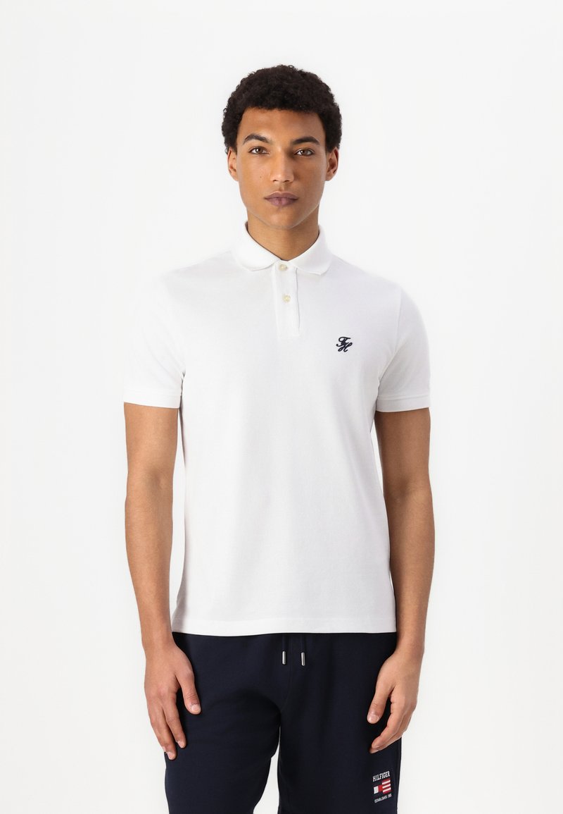 White polo shirt made from cotton, featuring a classic collar, two buttons, and a small black logo on the chest. Short sleeves.