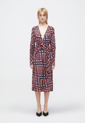 Femme debout, faisant face à l'avant, portant une robe enveloppante à manches longues et multicolore avec un décolleté en V profond et des chaussures noires à talons pointus.