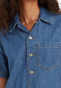 Denimová košile s límečkem, krátkými rukávy, světle modré barvy, kovovými knoflíky, náprsní kapsou a jemnými prošívacími detaily.