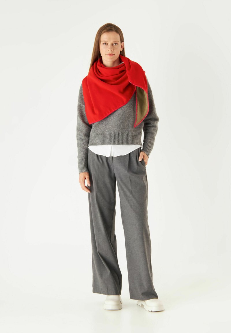 Grijze gebreide trui, witte blouse met kraag, grijze brede broek en een rode sjaal om de schouders gedrapeerd. Witte schoenen zichtbaar.