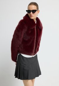 Burgundská umělá kožešinová bunda se zapínáním na zip a ribovanými lemy, kombinovaná s černou plisovanou sukní a bílým topem, stylizovaná s černými slunečními brýlemi ve tvaru kočičích očí.