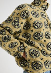 Pulover din fleece galben cu modele florale bleumarin, guler înalt, închidere cu fermoar pe jumătate și manșete elasticizate. Suprafață moale, texturată.