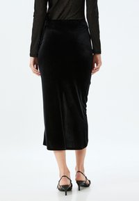Zwarte fluwelen midi-rok met een getailleerde silhouet, voorzien van een split aan de achterkant en een nette zoom. De gladde textuur complementeert het ontwerp.