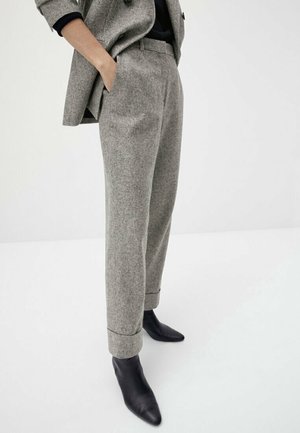 Person wearing gray checked tailored trousers with cuffs, black pointed ankle boots, and a matching blazer with hands in pockets.