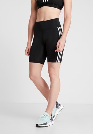 Frau trägt schwarze High-Waist-Sportshorts mit weißen Seitenstreifen und hellblauen Laufschuhen, steht vor einem einfarbigen Hintergrund.