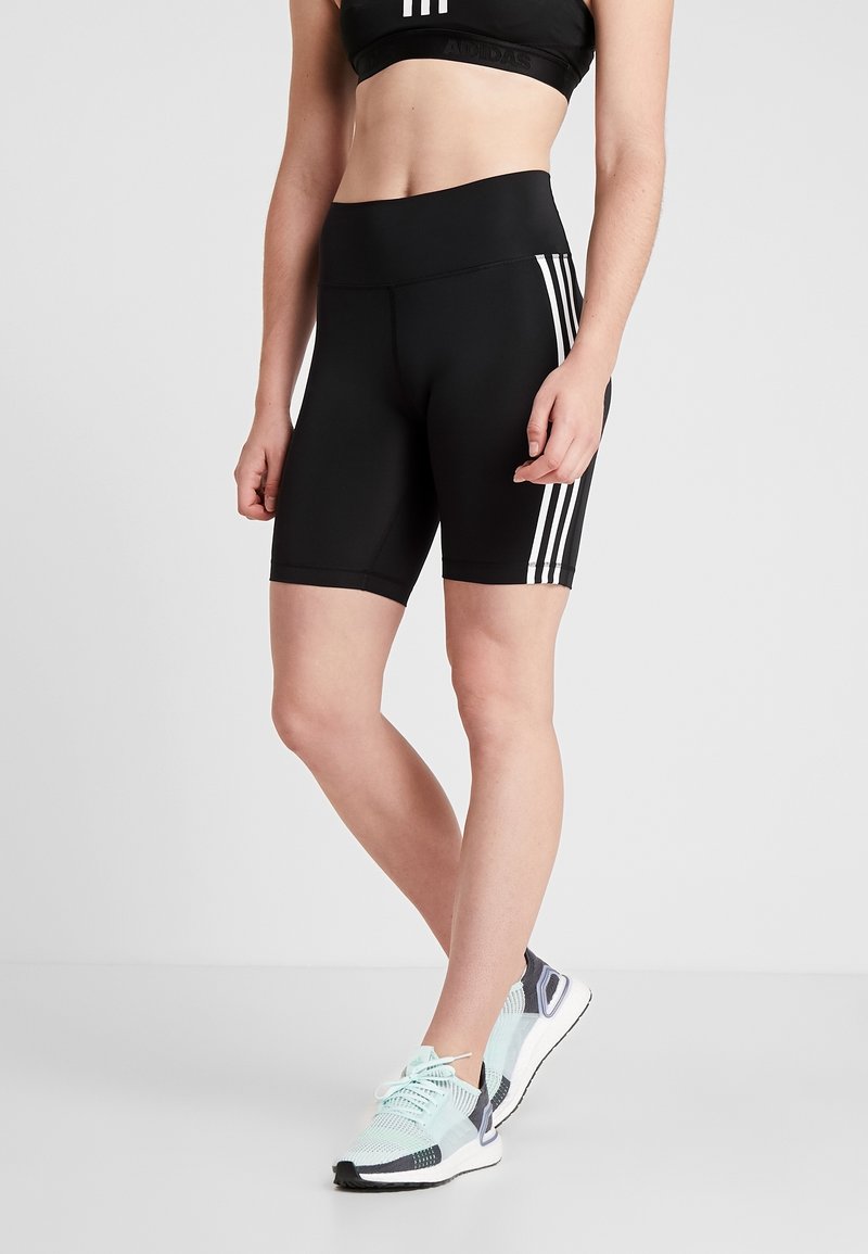 Femme portant un short de sport noir taille haute avec des bandes blanches sur les côtés et des chaussures de course bleu clair, debout devant un fond uni.
