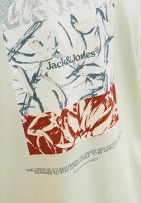 Világoszöld pamut póló, amely absztrakt virágmintákkal díszített grafikai tervezést mutat be kék és rozsdavörös színekben, valamint "Jack & Jones" logóval nyomtatva.