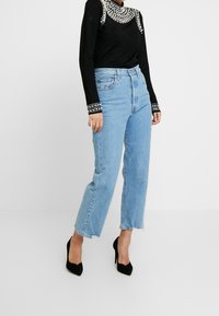 Schwarzes Langarmoberteil mit Nietenakzenten am Ausschnitt und an den Ärmeln, kombiniert mit hochgeschnittenen hellblauen Denim-Jeans mit fransigem Saum.