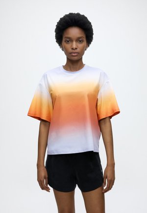 Žena s kratkom kovrčavom kosom nosi labavu majicu s gradijentom u bijeloj, žutoj i narančastoj boji u paru s crnim kratkim hlačama, stoji nasuprot bijele pozadine.