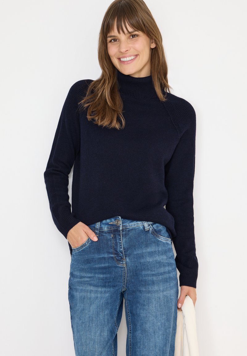 Pull à col roulé bleu marine avec des détails côtelés sur les épaules, assorti à un jean en denim bleu clair, avec une texture légère partout.