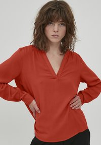 Blusa rossa a maniche lunghe con scollatura a V. Tessuto liscio e vestibilità rilassata. Dettagli notevoli includono spalle arr gathered e spacchi laterali.
