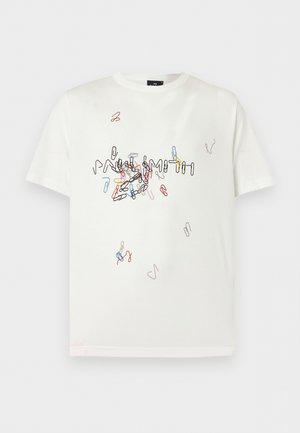 Homme aux cheveux bouclés portant un t-shirt blanc graphique, pantalon plissé gris, mocassins beige, debout avec une main dans la poche.