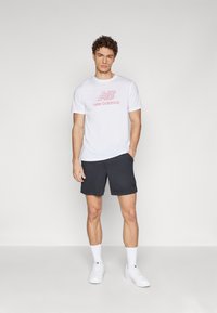 Balta kokvilnas T-kreklā ar rozā "New Balance" logotipu, savienots ar melniem sporta šortiem un baltiem sporta apaviem, uz vienkārša fona.