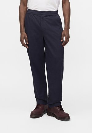 Homme portant un pantalon bleu marine foncé et des bottes marron foncé à lacets, présenté de la taille aux pieds sur un fond clair uni.