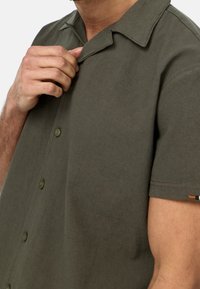 Camisa de manga corta de color verde oliva en un tejido liso. Cuenta con un cuello tipo camp, cierre con botones en la parte delantera y un pequeño bolsillo en el pecho.