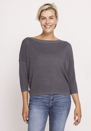 Smiling woman with short blonde hair wears a loose gray sweater and blue jeans, standing against a plain light background.