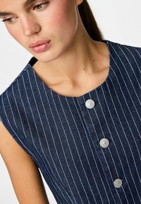 Vestido de mezclilla en azul oscuro con rayas verticales blancas, con escote redondeado y tres grandes botones grises en la parte delantera.