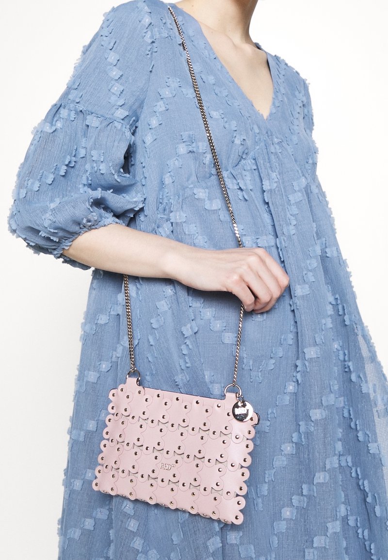 Femme tenant un sac à main rose à motif floral avec des clous métalliques et une bandoulière chaîne, portant une robe bleu clair texturée avec des manches bouffantes.