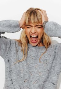 Pull en tricot gris clair avec un motif en vagues, doté d'un col rond et de manches trois-quarts. Cheveux longs et raides blonds encadrant le visage.