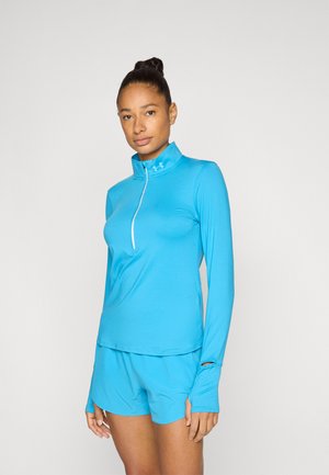 Vrouw draagt een felblauwe lange mouw sporttop met duimgaten en bijpassende shorts, staand tegen een effen witte achtergrond.