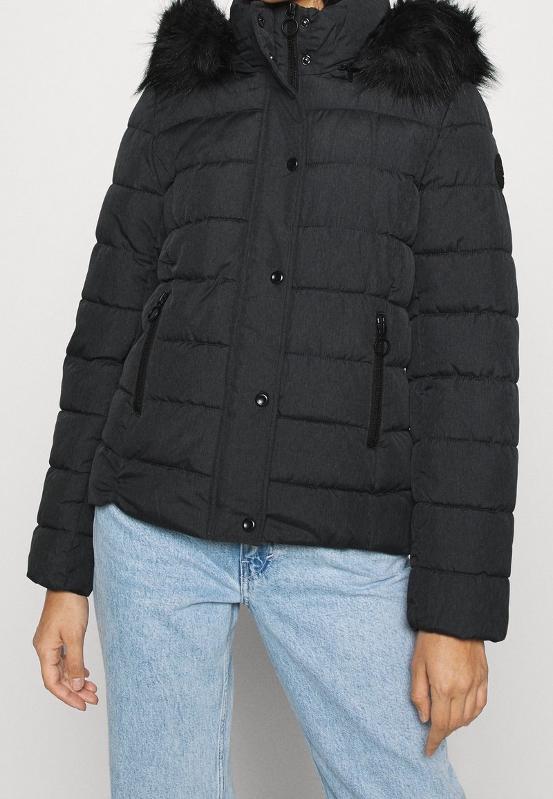Personne portant une veste d'hiver noire matelassée avec une capuche bordée de fourrure et des poches latérales zippées, associée à un jean bleu clair.