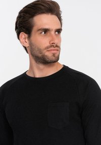 Zwarte geribbelde longsleeve shirt met een ronde hals en een klein borstzakje. De stof heeft een gladde textuur met contrasterende details op de mouwen.