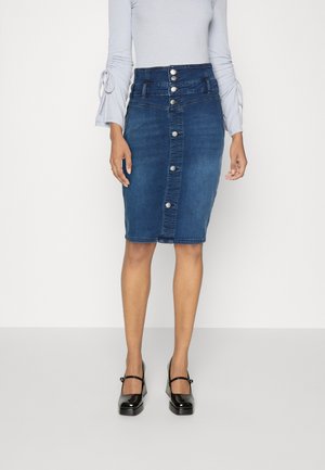 Frau trägt einen hochschnittigen Bleistiftrock aus Denim mit Knopfleiste, ein hellblaues langärmeliges Oberteil und schwarze Mary Jane Schuhe.