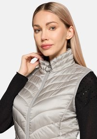Veste sans manches grise claire isolante avec un col montant, fermeture éclair et texture à coutures en chevrons ; portée sur un haut à manches longues noir avec des accents métalliques.