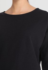 Schwarzes, strukturiertes Langarmshirt mit weitem Ausschnitt, das ein dezentes Webmuster und eine lockere Passform aufweist.