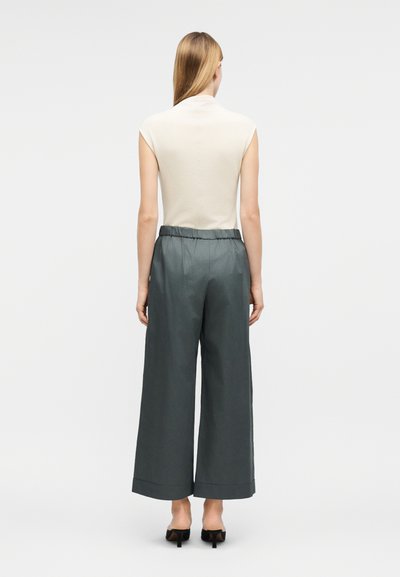 Γυναίκα με μακριά ξανθά μαλλιά που φοράει άσπρο τοπ χωρίς μανίκια, φαρδιά γκρι σκούρα παντελόνια και μαύρα παπούτσια με τακούνι, γυρισμένη με την πλάτη σε απλό φόντο.